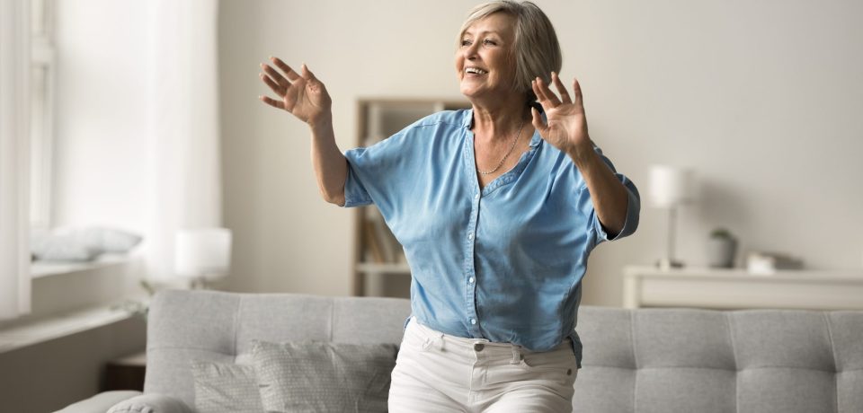 Smiling,Mature,Woman,Moves,To,Favourite,Song,In,Living,Room, Older adult woman performs the Timed Up and Go (TUG) Test to assess mobility, balance, and fall risk, demonstrating a functional movement assessment used by physical therapists.